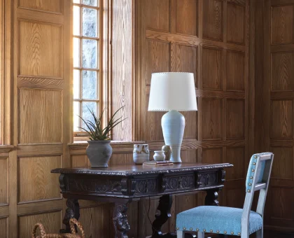 Traditional wood-paneled room with a carved dark wooden desk, a ceramic lamp, a plant, and a wicker basket at the base of the desk.