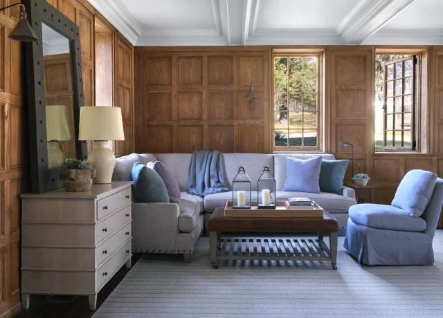 Wood-paneled living room with a gray sofa, blue pillows, and a light throw; white coffered ceiling and large windows letting in natural light