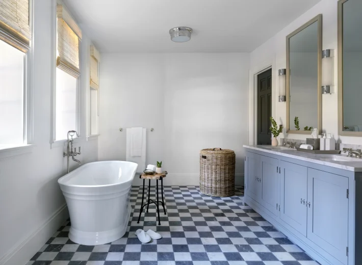 White freestanding bathtub beside a black-and-white checkered floor in a bright bathroom with a wicker laundry basket nearby