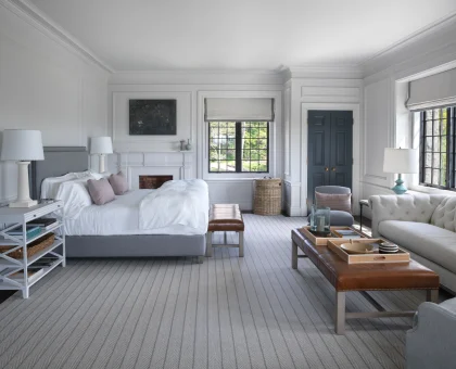 Bright bedroom with white paneled walls, two beds, and soft gray decor; large windows and a blue double door visible in the background.
