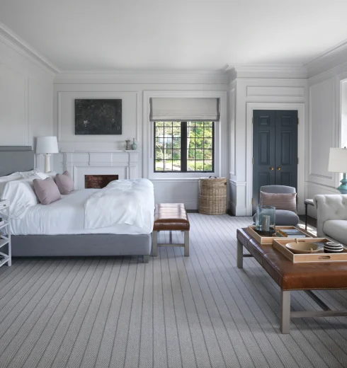 Bright bedroom with white paneled walls, two beds, and soft gray decor; large windows and a blue double door visible in the background.