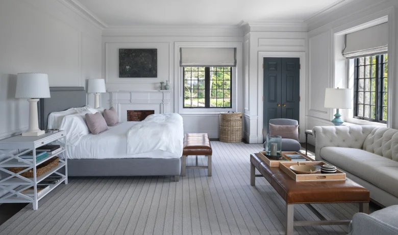Bright bedroom with white paneled walls, two beds, and soft gray decor; large windows and a blue double door visible in the background.