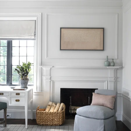 Bright living room with white paneled walls, a fireplace, and a beige abstract artwork above the mantel; a gray upholstered chair and a woven basket of logs nearby, plus a window with a blind and a potted plant.