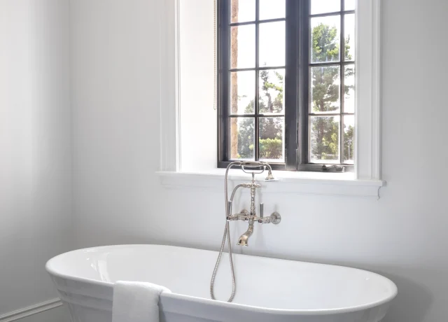 White freestanding bathtub under a large grid-window with a beige Roman shade; black-and-white checkered floor visible in a bright bathroom.