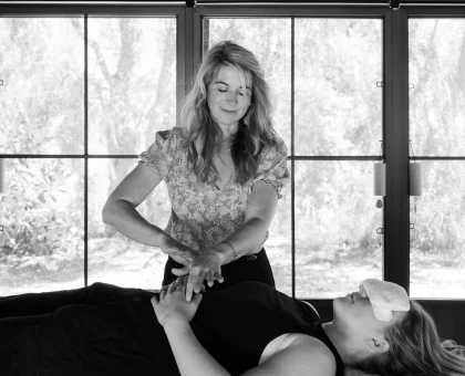 Therapist giving a back massage to a client lying on a table in a bright room with large windows.