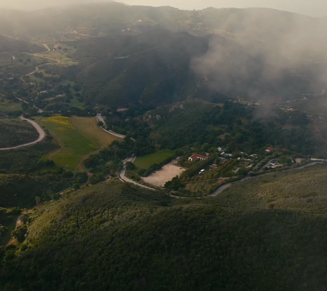 Aerial view of a winding road through a green, hilly valley with scattered houses and fields, under hazy light with smoke in the distance.