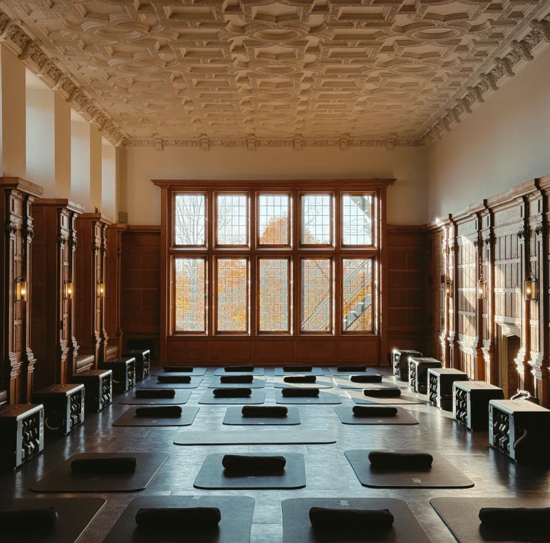Historic-style meditation hall with multiple yoga mats arranged in neat rows before a large paneled window wall.