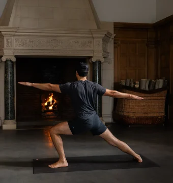 Person in a dark shirt and shorts performing Warrior II yoga pose on a mat near a lit fireplace indoors. No depiction of faces required for accessibility.