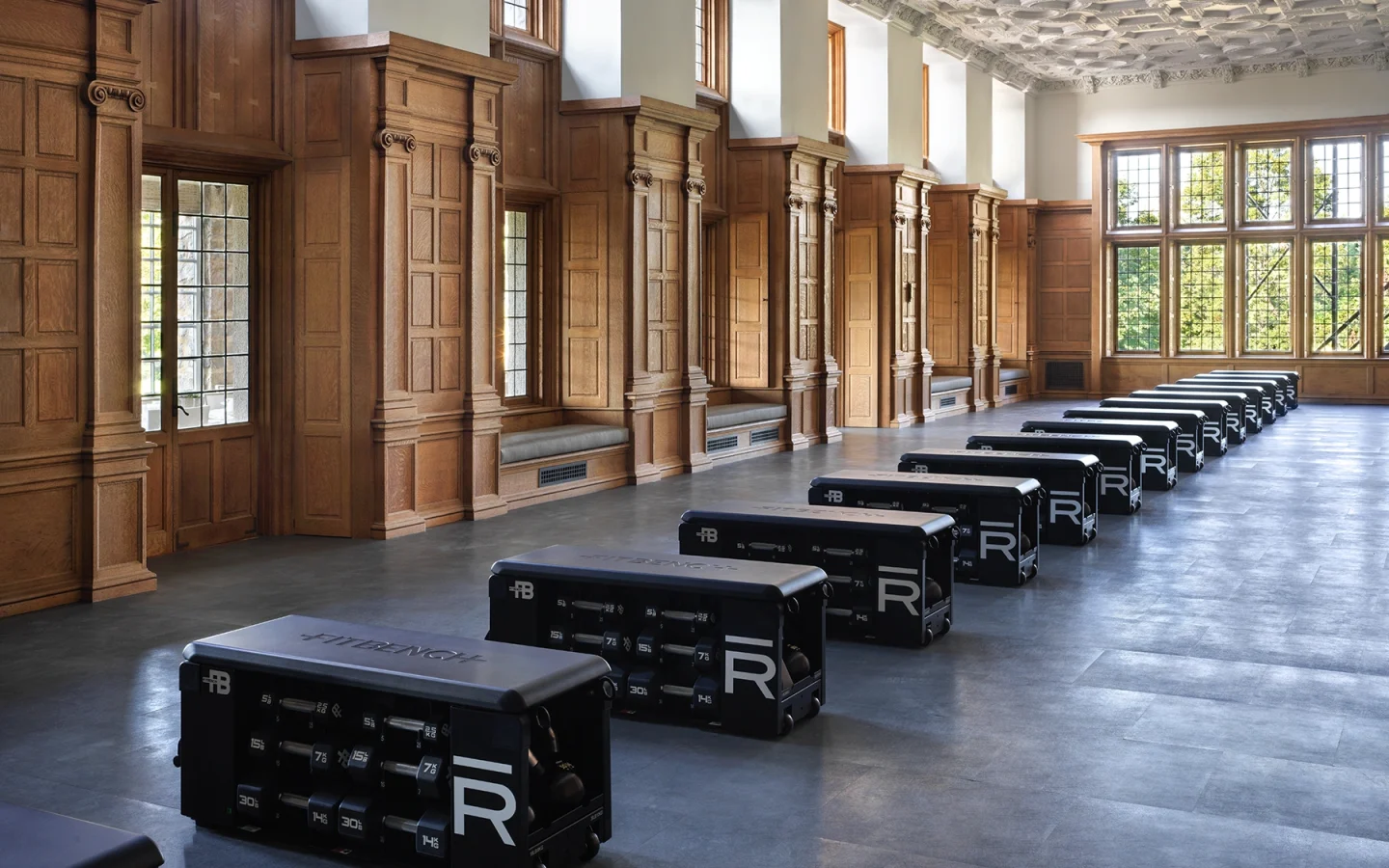 Historic-style gym with wood-paneled walls, tall windows, and rows of black reformer machines along the floor.