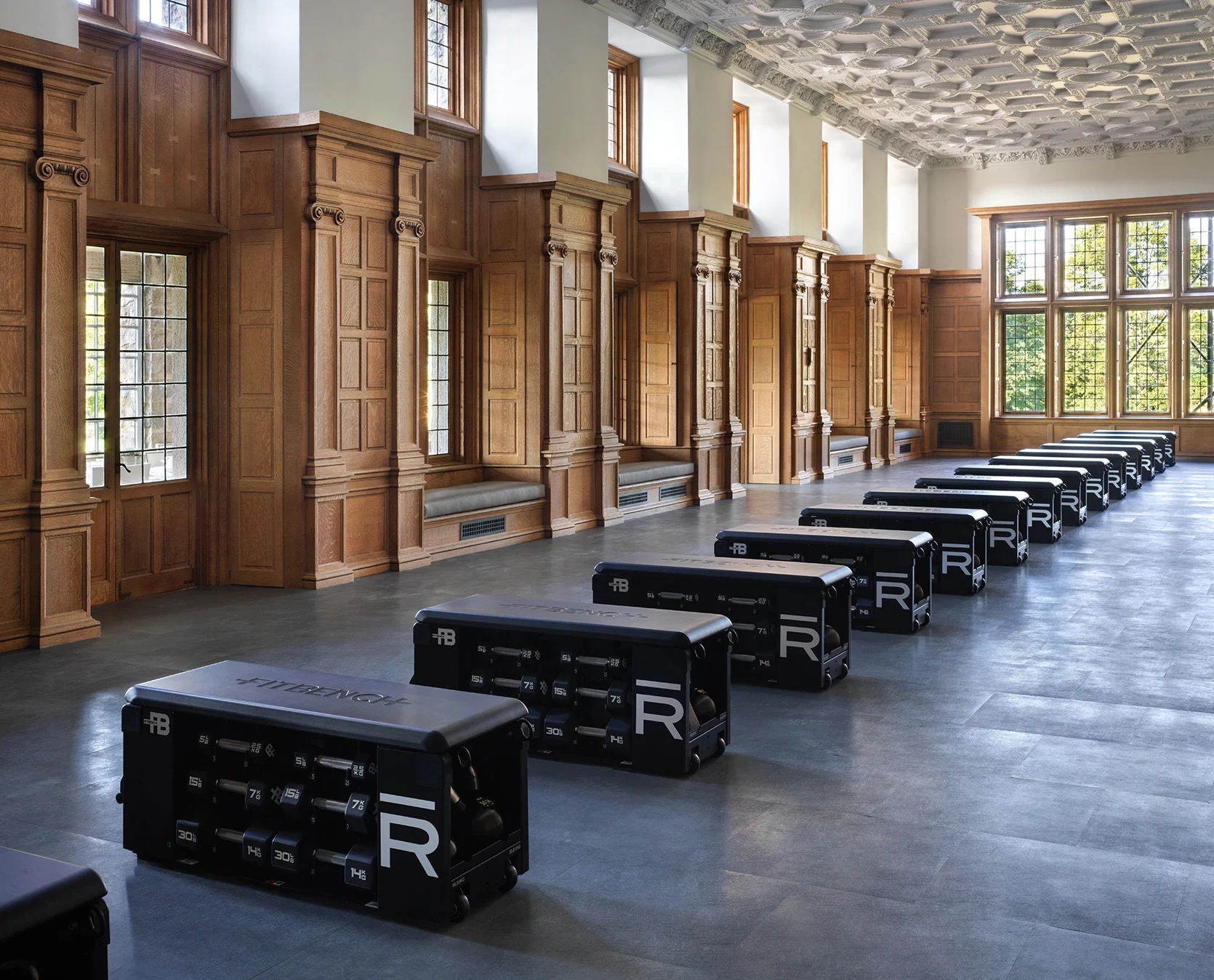 Historic-style gym with wood-paneled walls, tall windows, and rows of black reformer machines along the floor.