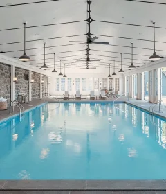 Indoor pool area with stone walls, bright blue water, and seating at the far end under a high, white ceiling with fans and pendant lights