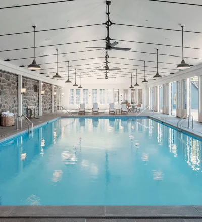 Indoor pool area with stone walls, bright blue water, and seating at the far end under a high, white ceiling with fans and pendant lights