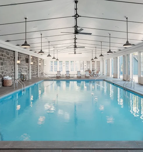 Indoor pool area with stone walls, bright blue water, and seating at the far end under a high, white ceiling with fans and pendant lights
