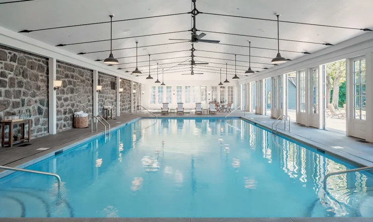 Indoor pool area with stone walls, bright blue water, and seating at the far end under a high, white ceiling with fans and pendant lights