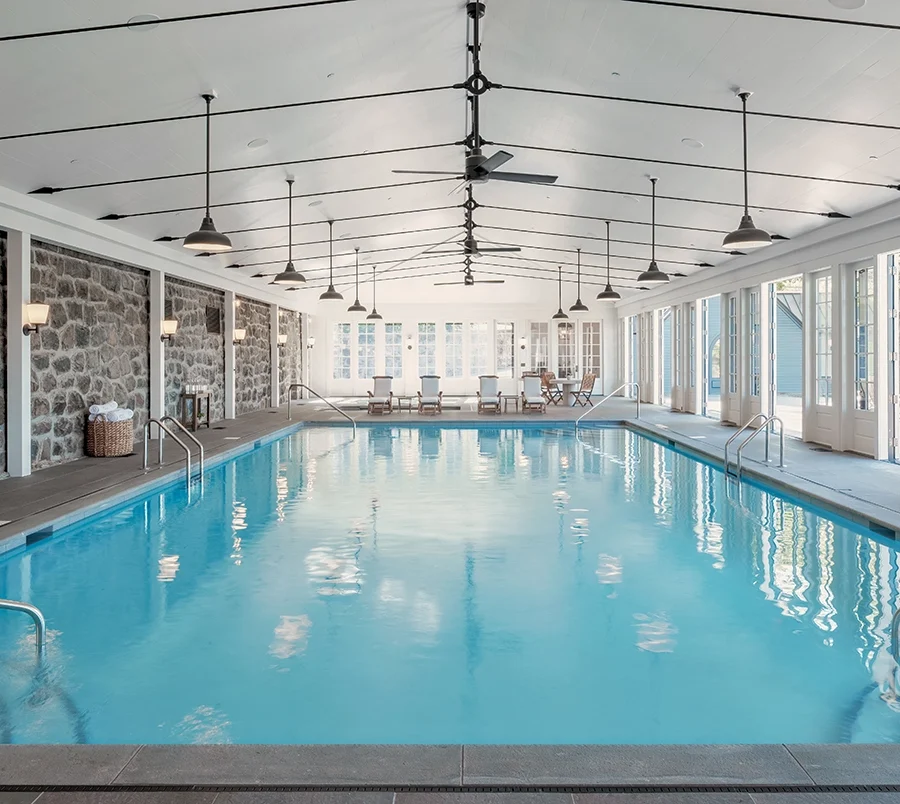 Indoor pool area with stone walls, bright blue water, and seating at the far end under a high, white ceiling with fans and pendant lights
