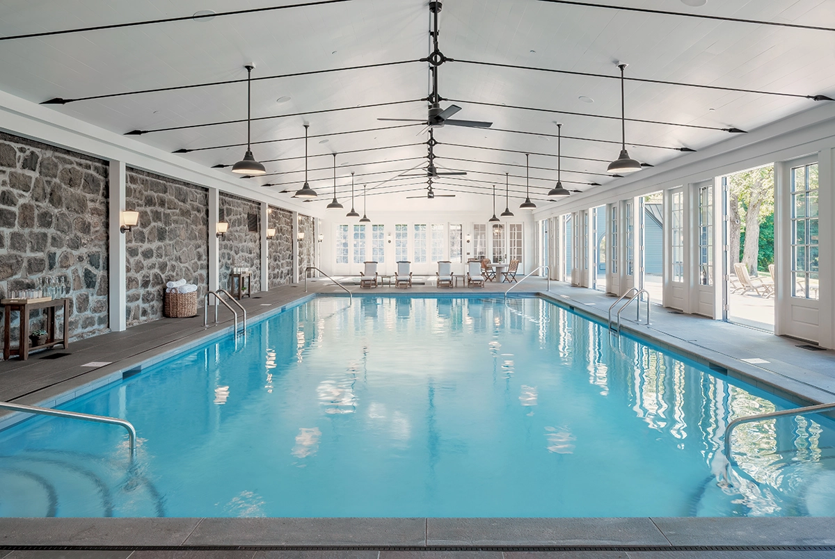 Indoor pool area with stone walls, bright blue water, and seating at the far end under a high, white ceiling with fans and pendant lights