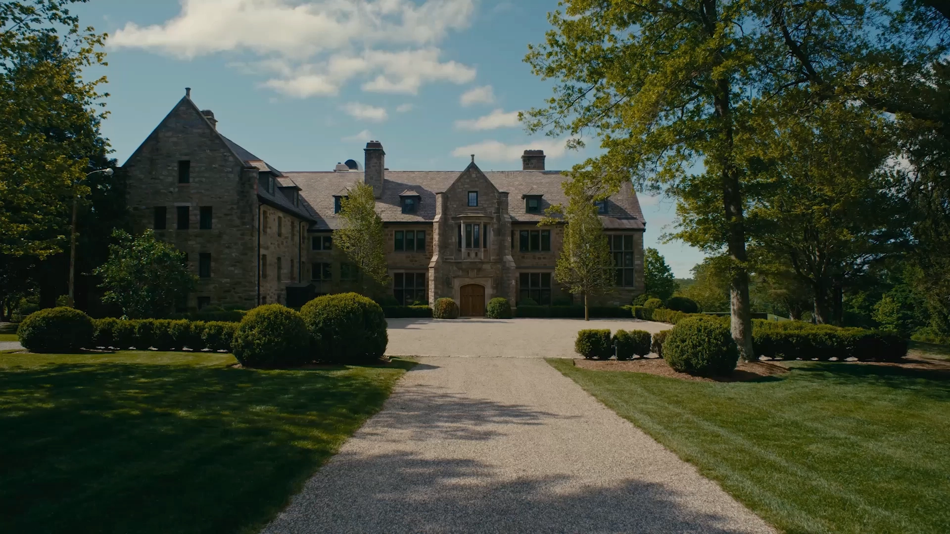 Front view of a historic stone mansion with a central arched wooden door, flanked by tall windows and manicured shrubs on a sunny day.