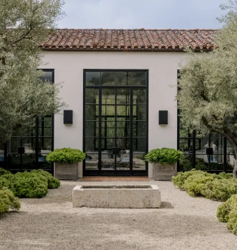 Modern building facade with large black-framed glass doors, beige walls, and a red terracotta-tiled roof, framed by olive trees.