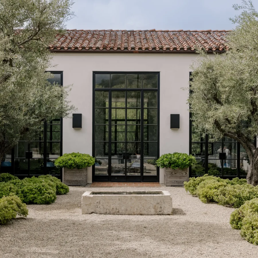 Modern building facade with large black-framed glass doors, beige walls, and a red terracotta-tiled roof, framed by olive trees.