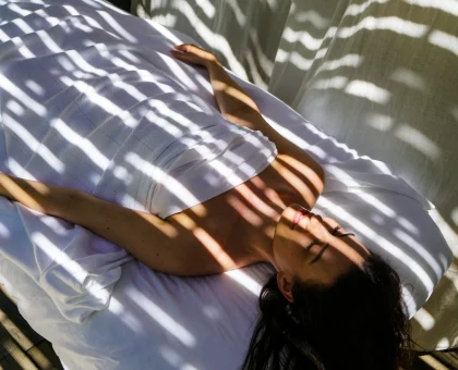 A person resting on a white blanket, shadows from window blinds create striped patterns across the bed and their body.
