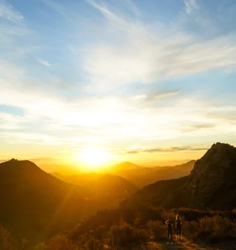 Sunset over rugged mountains with a warm golden glow, two hikers on a trail in the foreground.