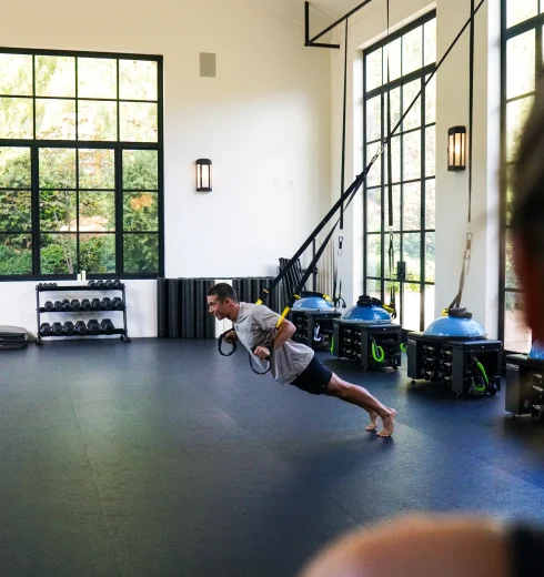 Man performing a suspended push-up with resistance straps in a bright gym with large windows and workout equipment along the wall