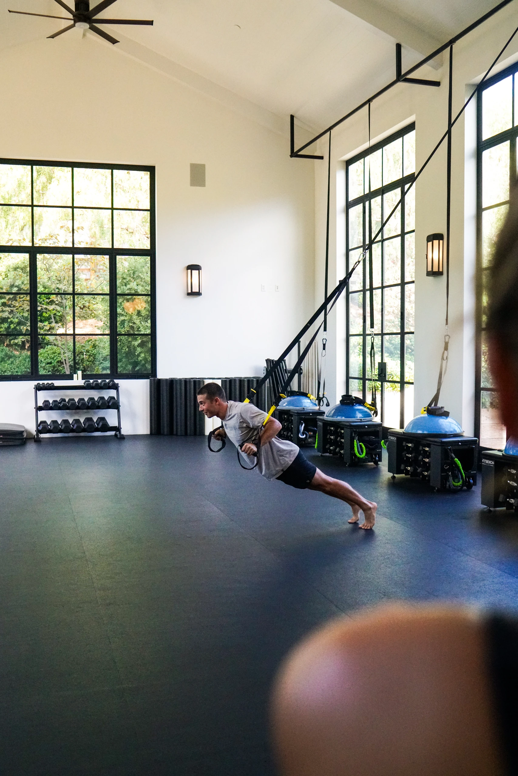 Man performing a suspended push-up with resistance straps in a bright gym with large windows and workout equipment along the wall