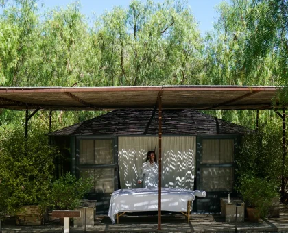 Outdoor wooden hut with a bed set up under a corrugated roof, surrounded by green trees; person in a white robe stands in the doorway.