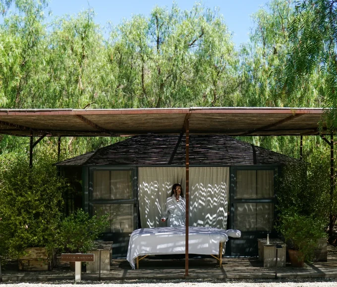 Outdoor wooden hut with a bed set up under a corrugated roof, surrounded by green trees; person in a white robe stands in the doorway.
