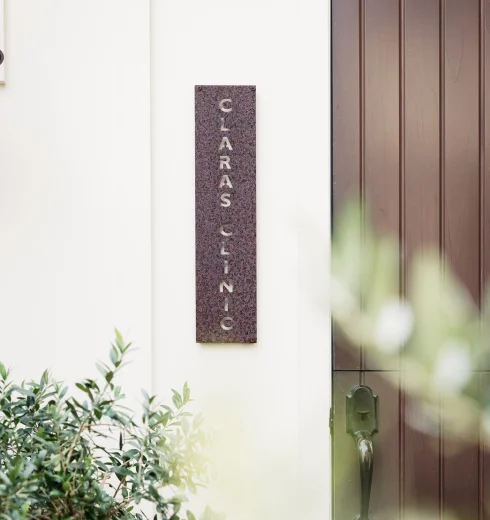 Vertical rust-colored metal sign with vertically arranged letters mounted on a pale wall beside a dark wooden door.