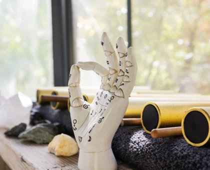 Ceramic hand model showing acupuncture pressure points on a wooden workbench with small stones and tools nearby by a windowed background.