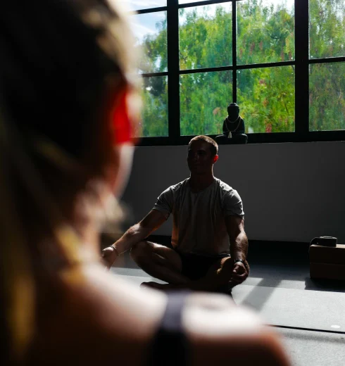 Meditating person sits cross-legged on the floor in a sunlit studio, large grid window behind them with a small Buddha statue on the sill.