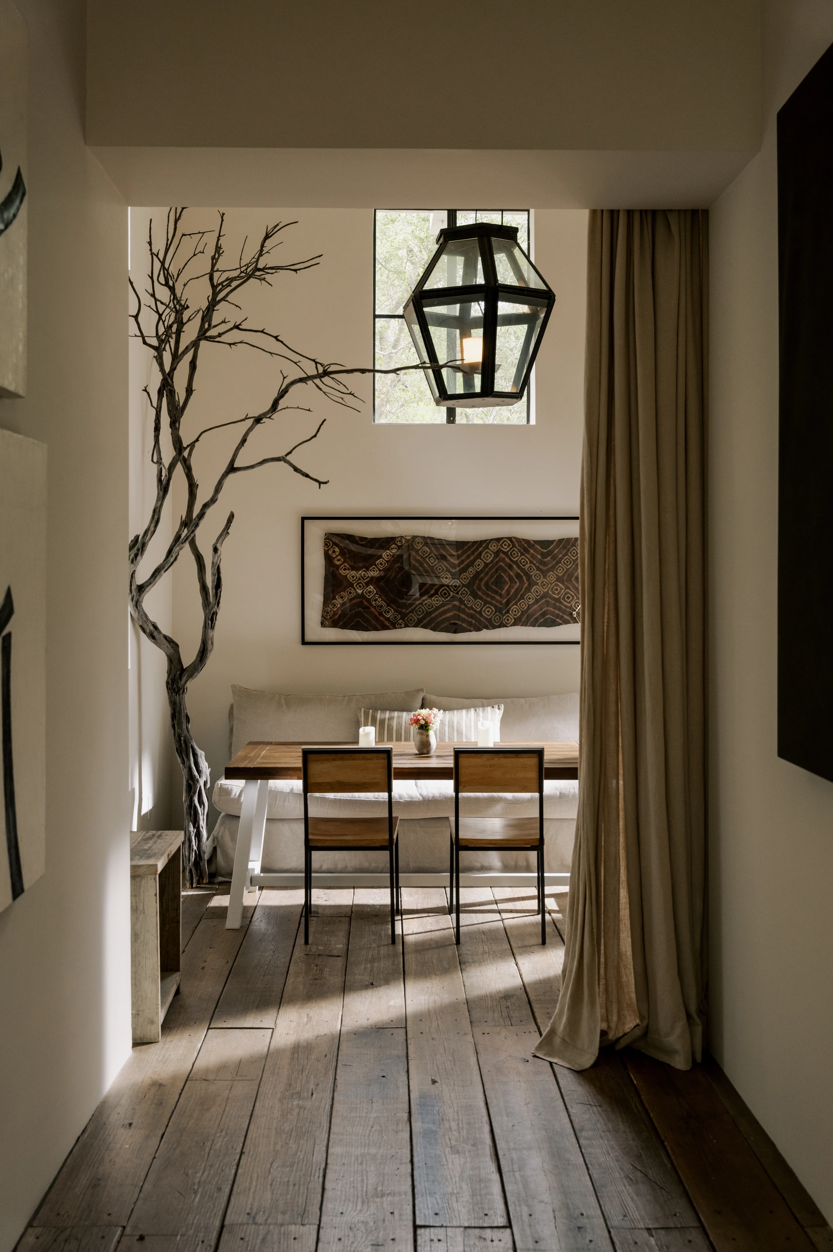 Dining nook with wooden table and four chairs, a geometric hanging lantern, and a bare-tree sculpture near a window.