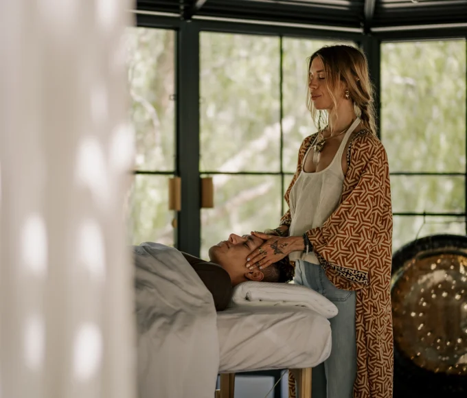 Massage therapist stands beside a client lying on a padded table, gently supporting the client's head in a bright, modern room with large windows.