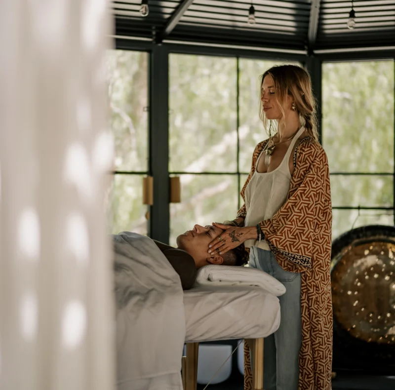 Massage therapist stands beside a client lying on a padded table, gently supporting the client's head in a bright, modern room with large windows.