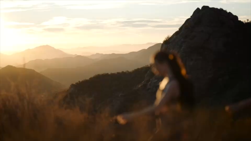 Golden hour mountain landscape at sunset with a blurred person in the foreground on a hillside.