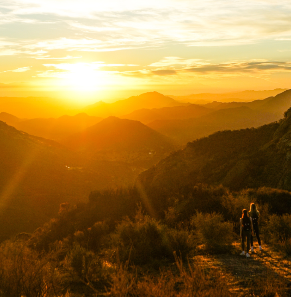 Sunset over rolling mountains with two hikers standing on a trail, bathed in warm orange light.