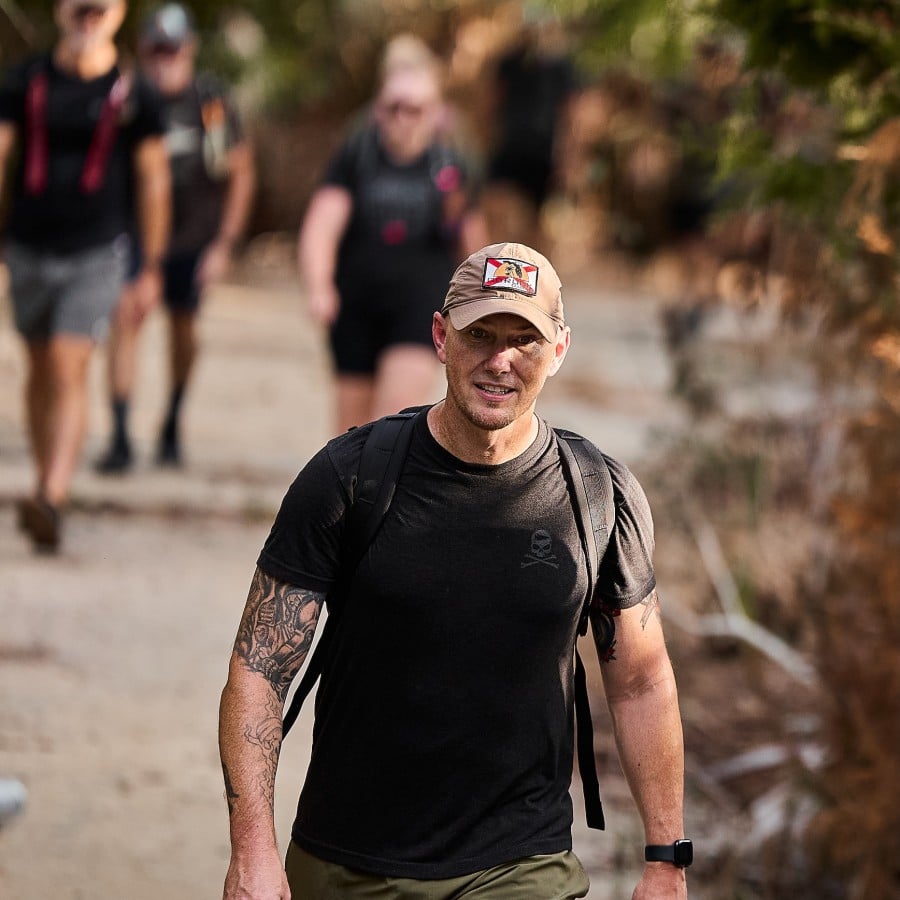 Man in a black t-shirt with a backpack walks toward the camera on a sandy trail, wearing a beige cap with a patch and showing a tattooed forearm; blurred hikers in the background.