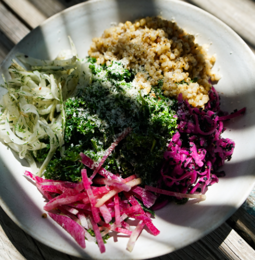 White plate with a grain bowl and colorful toppings: cucumber ribbons, dill-scented cabbage, pickled red onions, chopped herbs, and crumbled grains on top.