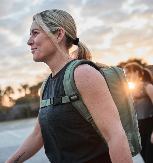 Side profile of a woman with blonde hair in a ponytail wearing a green backpack outdoors at sunset.