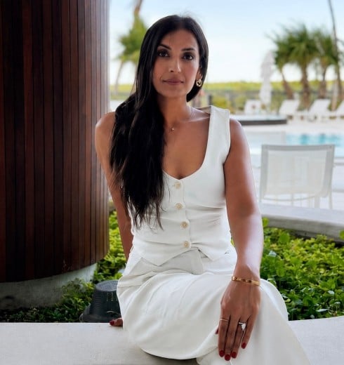 Woman with long dark hair in a white sleeveless button-front dress sits by a poolside, with greenery and palm trees in the background.