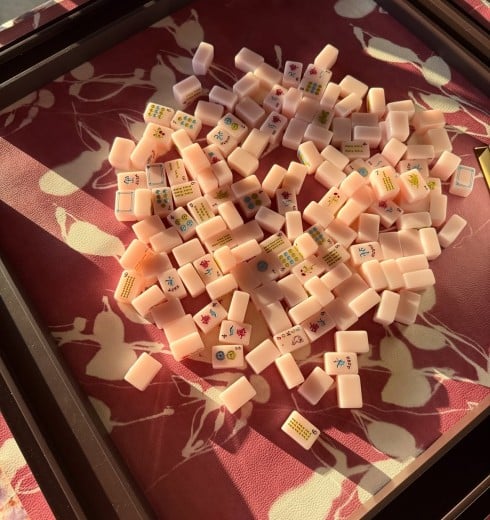 Pile of small pale pink Mahjong tiles with Chinese characters and symbols scattered in a shallow tray on a patterned red surface.