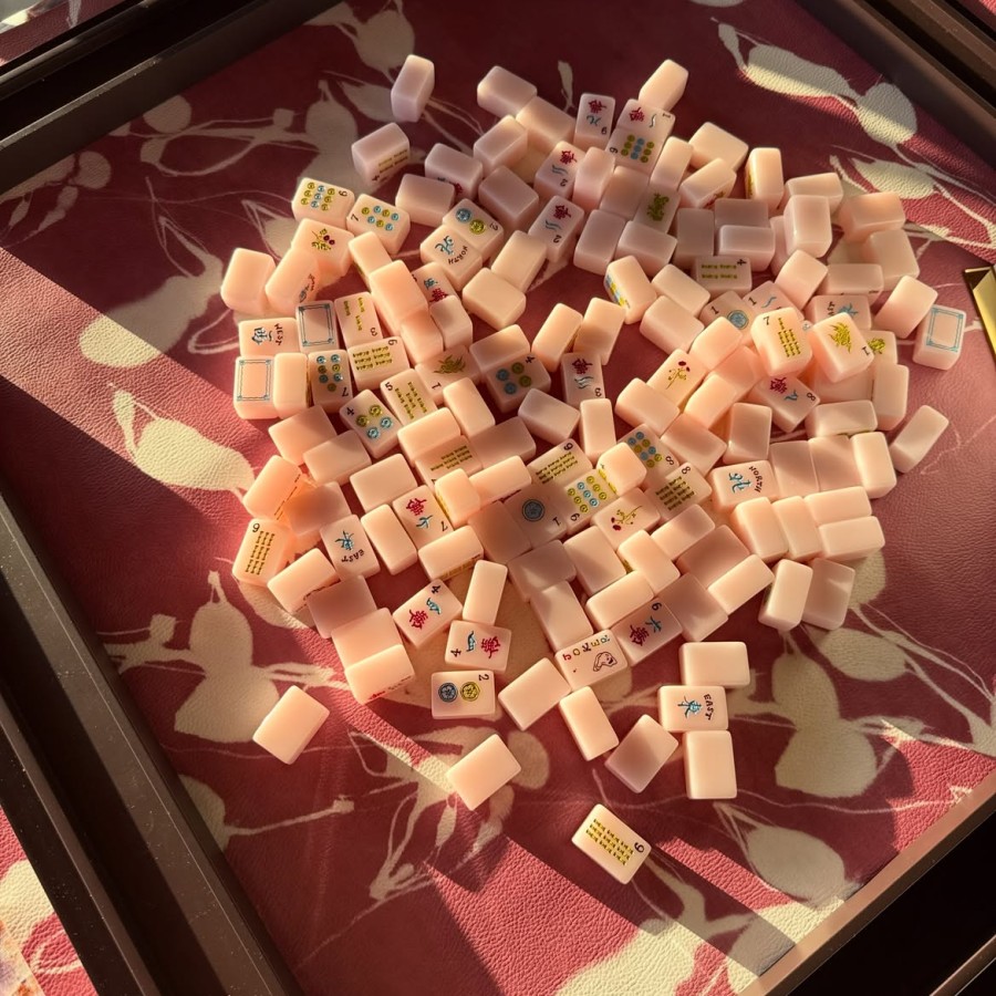 Pile of small pale pink Mahjong tiles with Chinese characters and symbols scattered in a shallow tray on a patterned red surface.