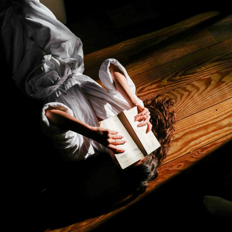 Person in a white robe lying on a wooden floor, reading an open book with dramatic lighting.