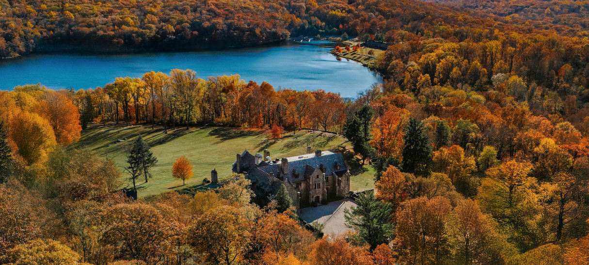 Stone mansion tucked among vibrant autumn trees beside a calm blue lake (aerial view).”,