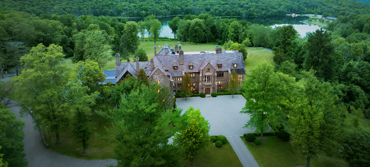 Aerial view of a large stone mansion surrounded by trees, with a gravel drive and manicured lawn. A lake is visible in the distance.