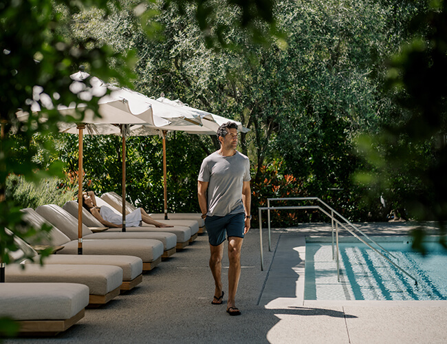 Man walking along a pool with lounge chairs and patio umbrellas nearby