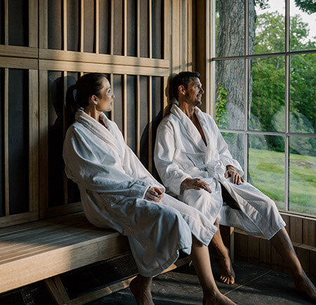 Couple sitting in a sauna together in white robes looking through an outside window